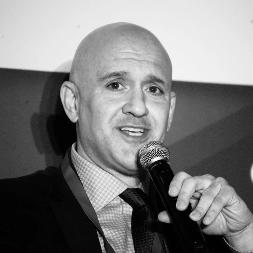 A bald man in a suit speaking into a microphone at an event.