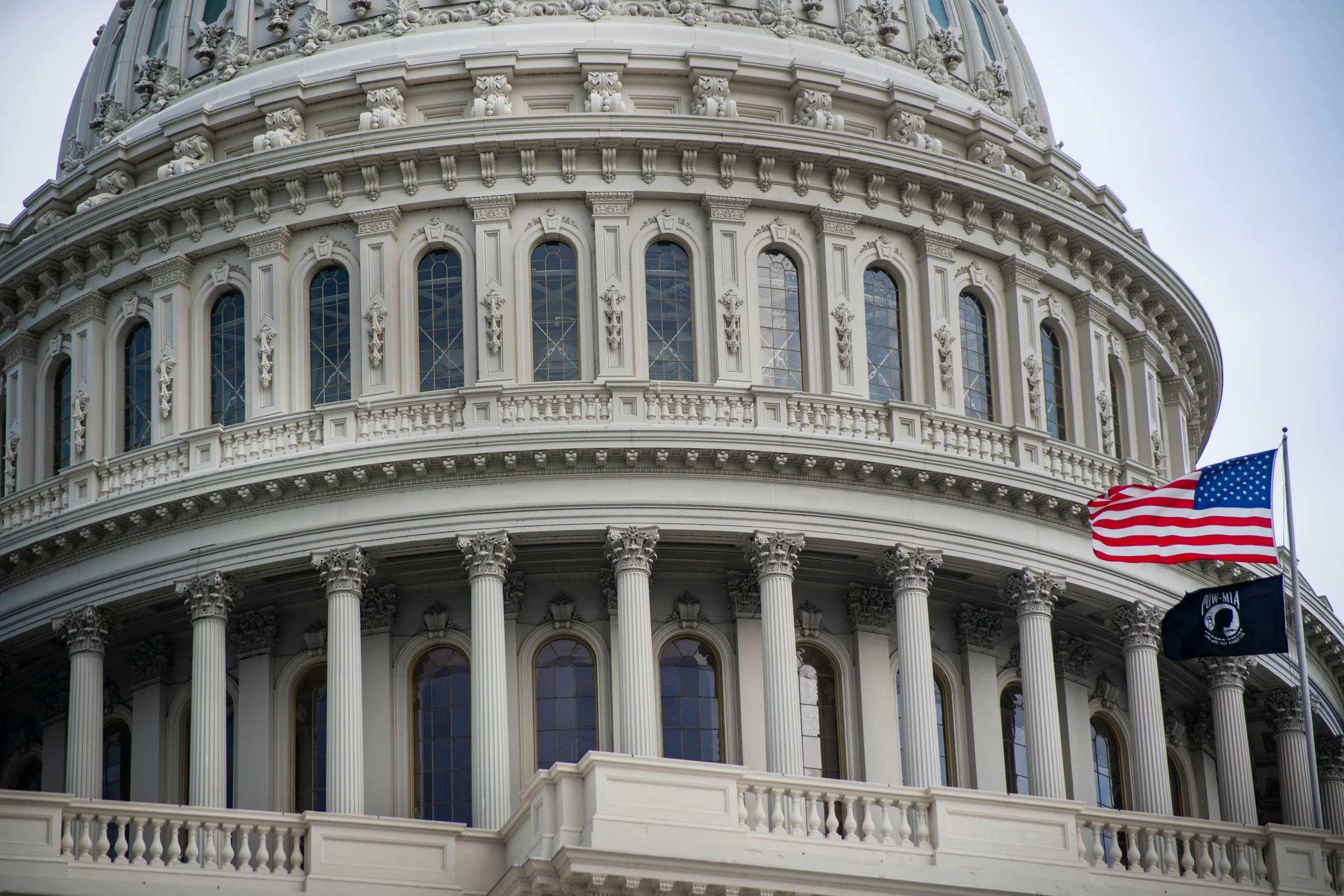 Close-up of the United States Capitol building's white ornate facade with columns, arched windows, and decorative details. American flags are flying in the foreground.