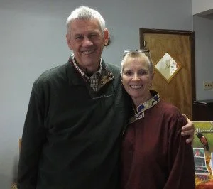 A man and woman standing together indoors, smiling at the camera, with a wooden door behind them.