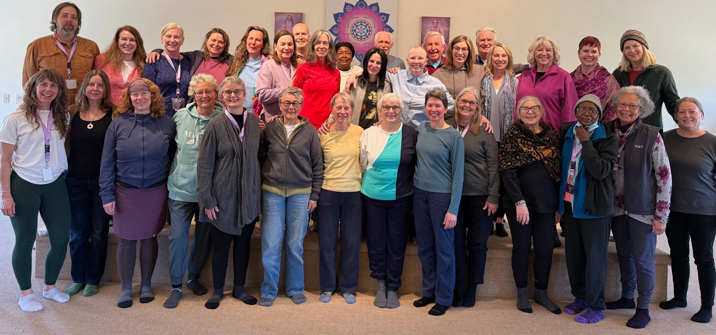 Group of smiling people in casual clothing standing on a stage inside a room with beige walls and religious artwork in the background.