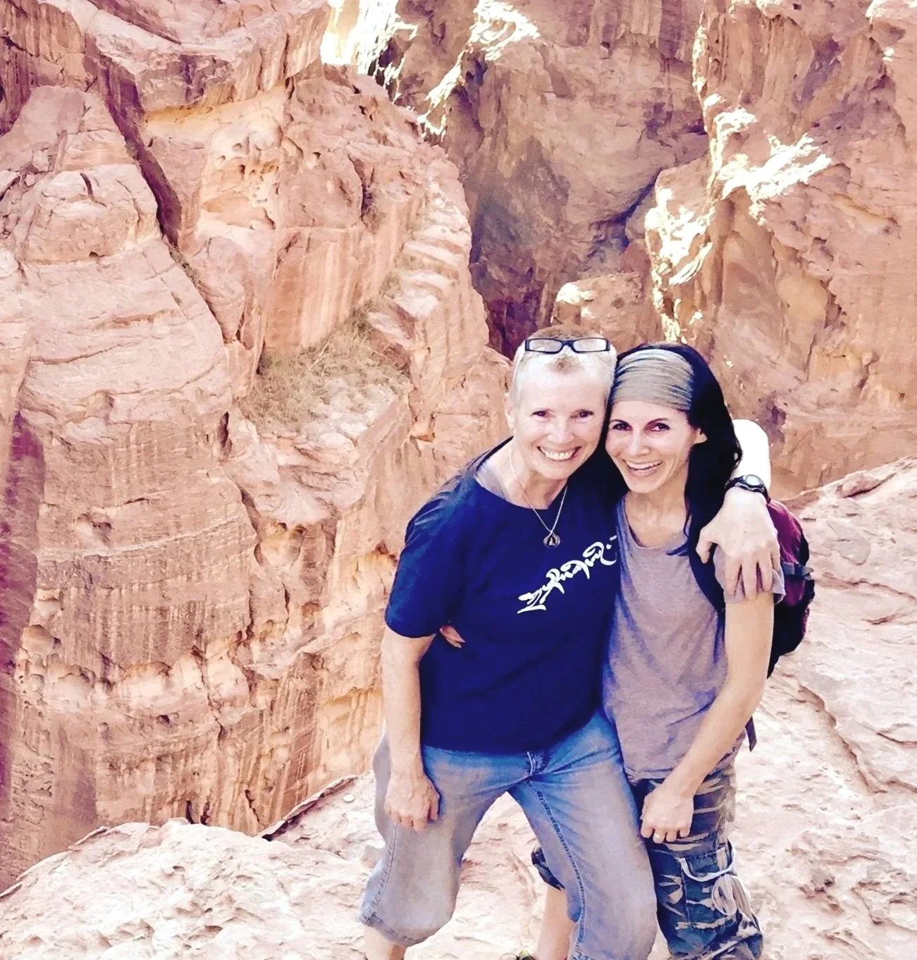 Two women standing close together in a rocky canyon, smiling at the camera with arms around each other, wearing casual outdoor clothing.