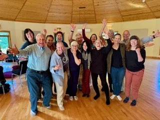 A woman in white clothing standing on stage with arms raised and eyes closed, speaking passionately. There are candles on a table and a flower arrangement behind her.