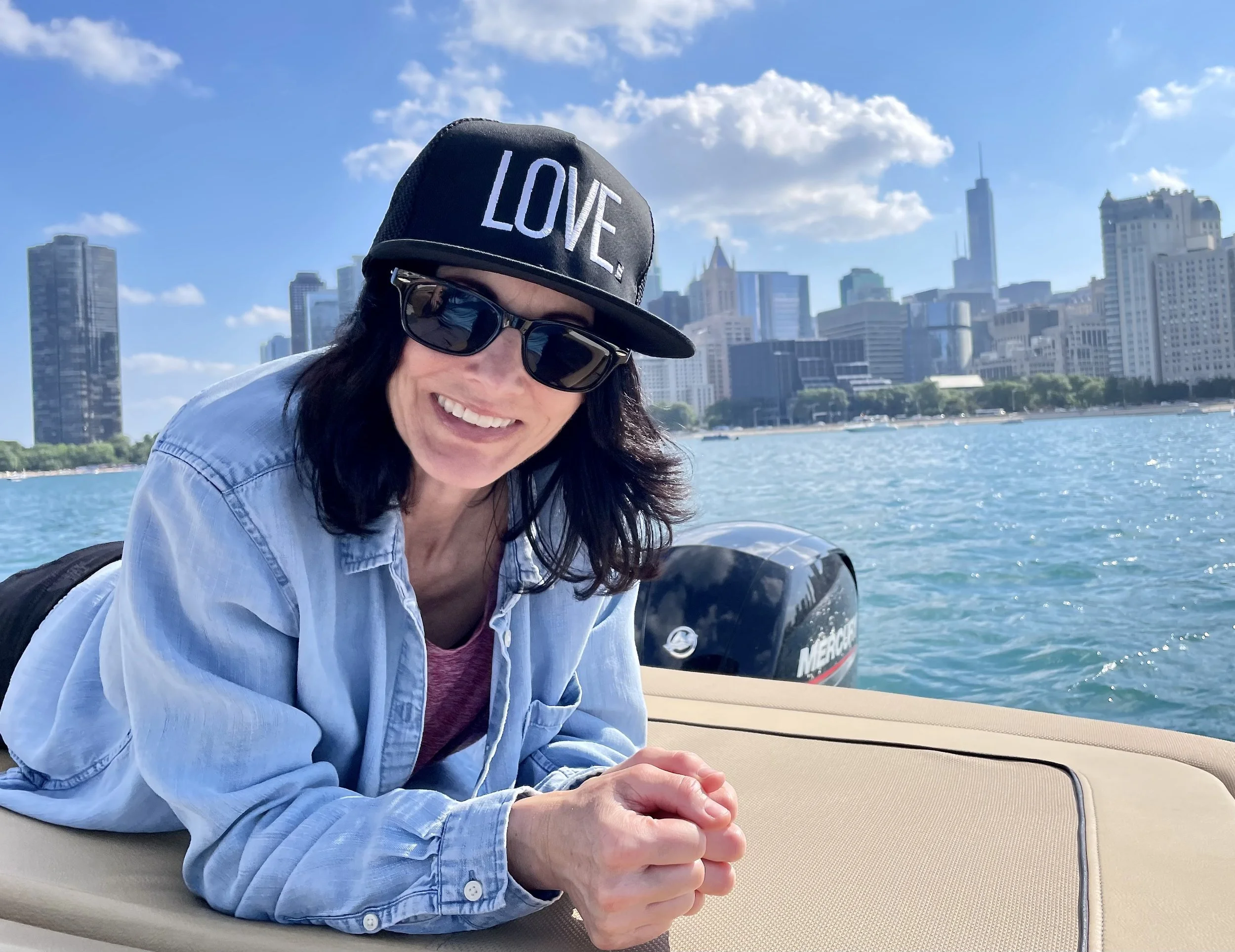 Smiling woman in sunglasses and a black cap with the word 'LOVE' on it, lying on a boat with a city skyline in the background on a sunny day.