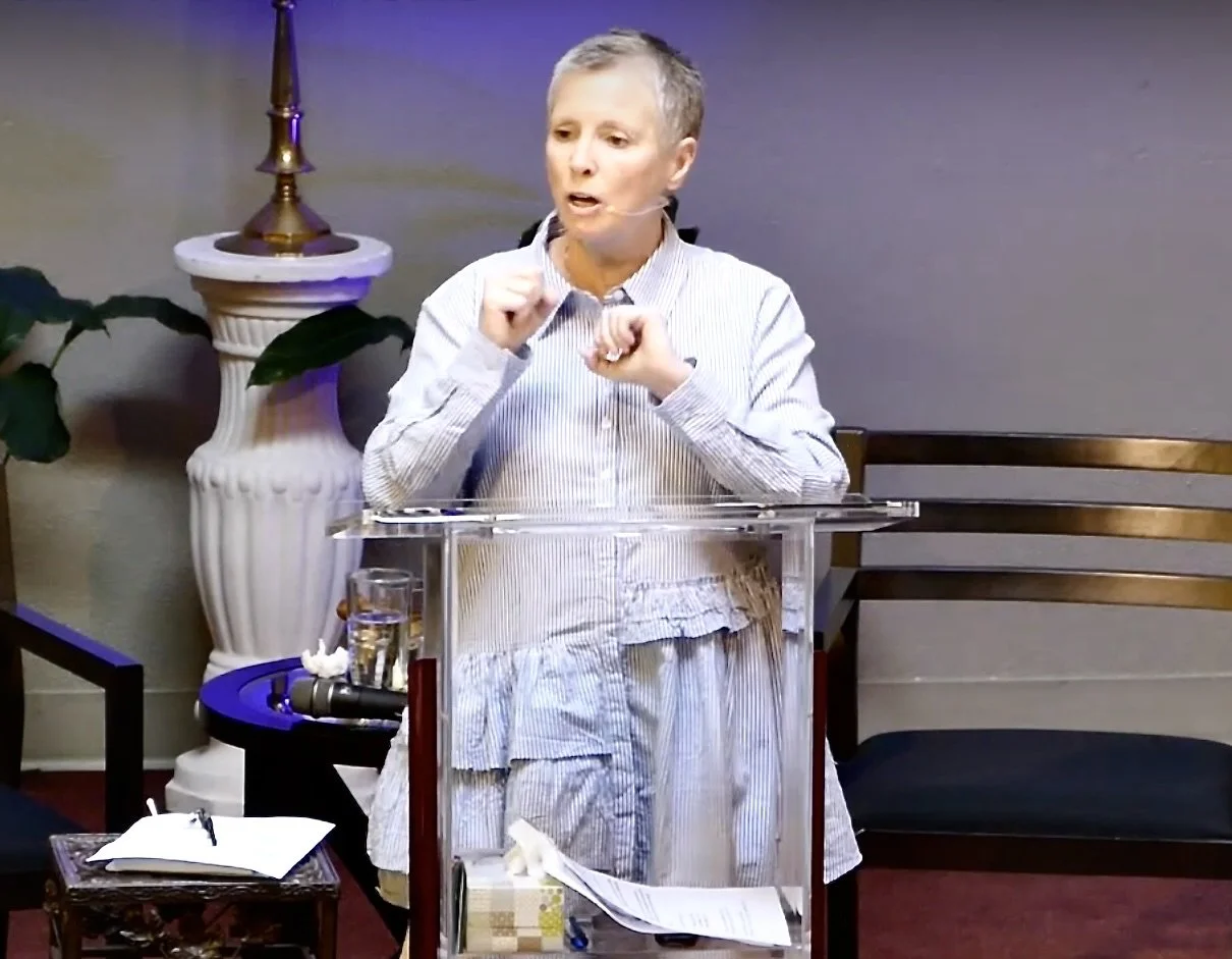 A woman with short gray hair wearing a striped dress standing behind a transparent podium, speaking passionately with clenched fists, a glass of water, and papers on a small table nearby, and a large decorative plant in the background.