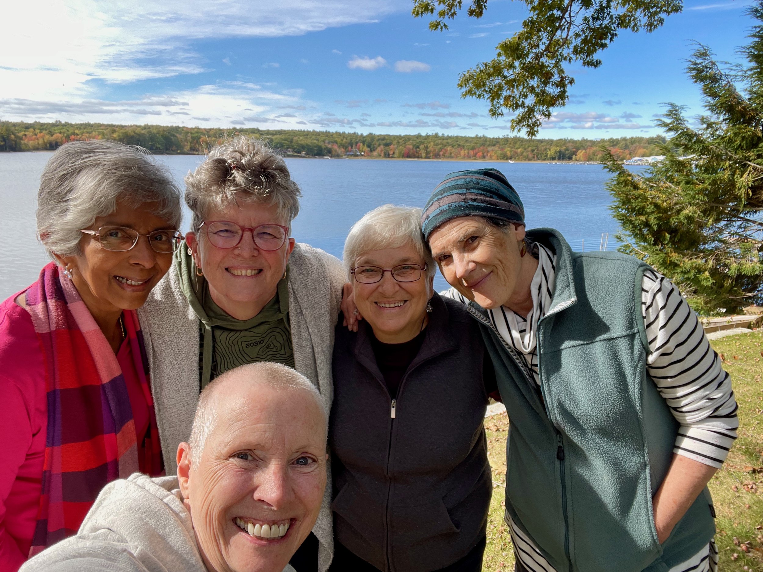 Group of five women smiling outdoors near a lake with trees and sky in the background.