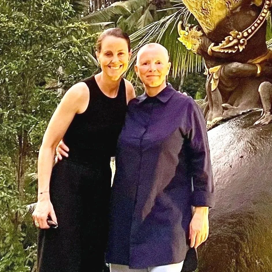 Two women standing outdoors next to a decorative sculpture of a frog with yellow flowers, surrounded by green foliage.