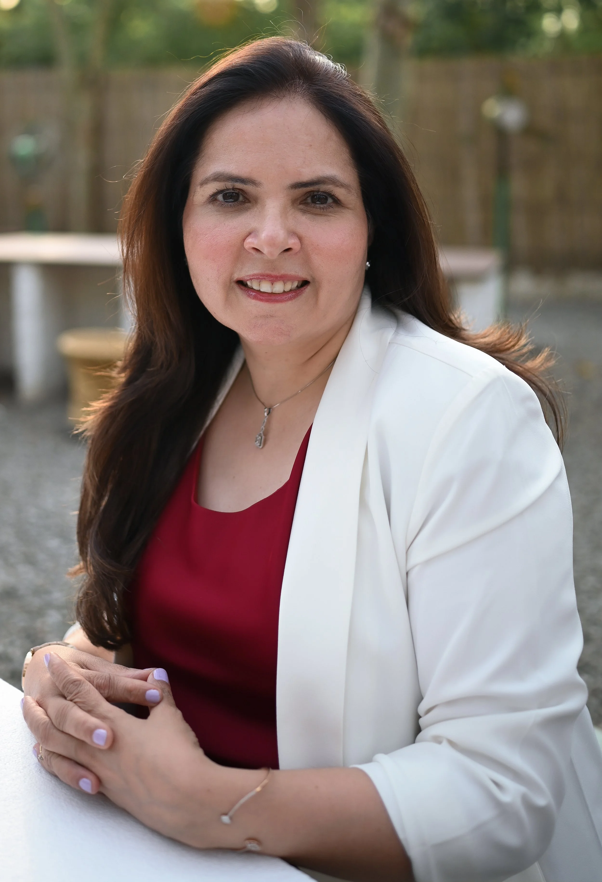 A woman with long dark hair wearing a white blazer over a red top, sitting outdoors with a blurred background of trees and outdoor furniture, smiling at the camera.