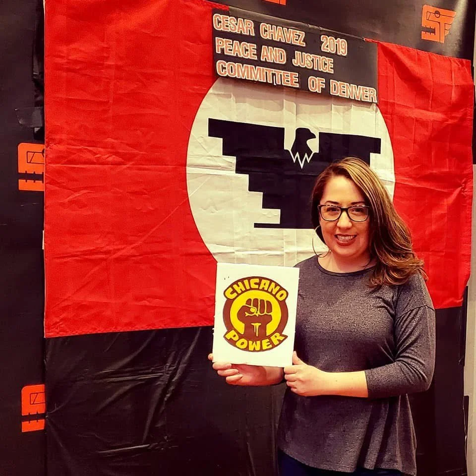 Justine Sandoval with glasses and long brown hair holding a sign that reads "Chicano Power" with a raised fist emblem. In front of a banner with a black eagle design and text referencing Cesar Chavez, 2019, Peace and Justice Committee of Denver.