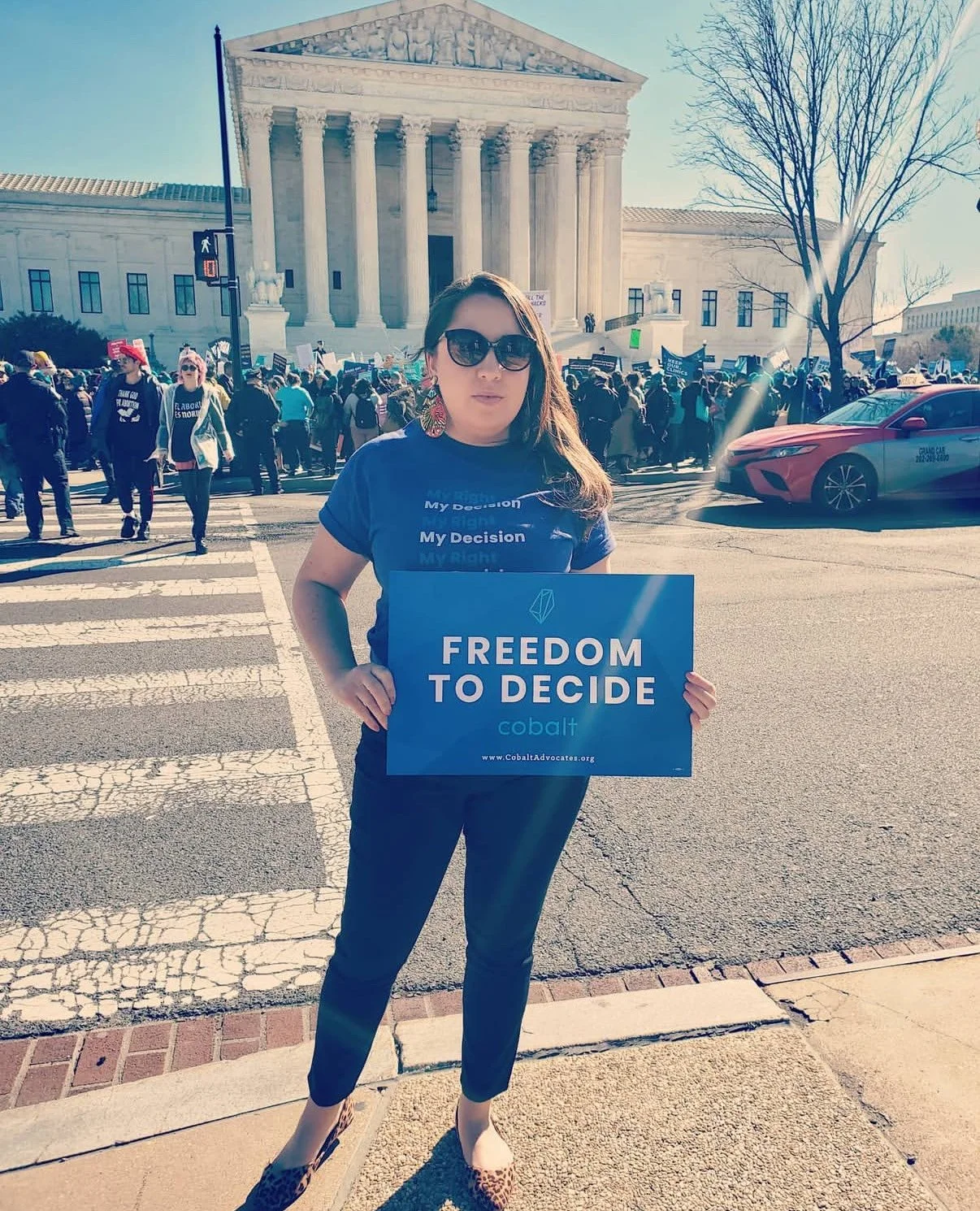 Justine Sandoval holding a blue sign that says 'FREEDOM TO DECIDE,' standing in front of the U.S. Supreme Court building during a protest or rally, with a crowd in the background.