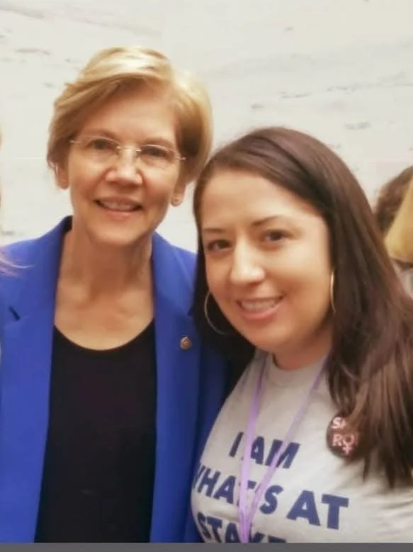 Elizabeth Warren and Justine Sandoval smiling for a photo indoors, one wearing a blue blazer and glasses, the other with long dark hair and a gray T-shirt with purple text.