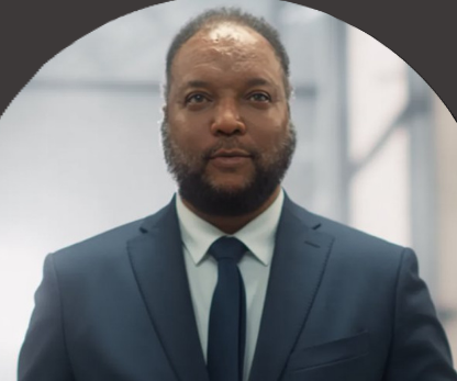 A man with dark skin, short hair, and a beard, wearing a navy blue suit and tie, standing against a blurred indoor background.