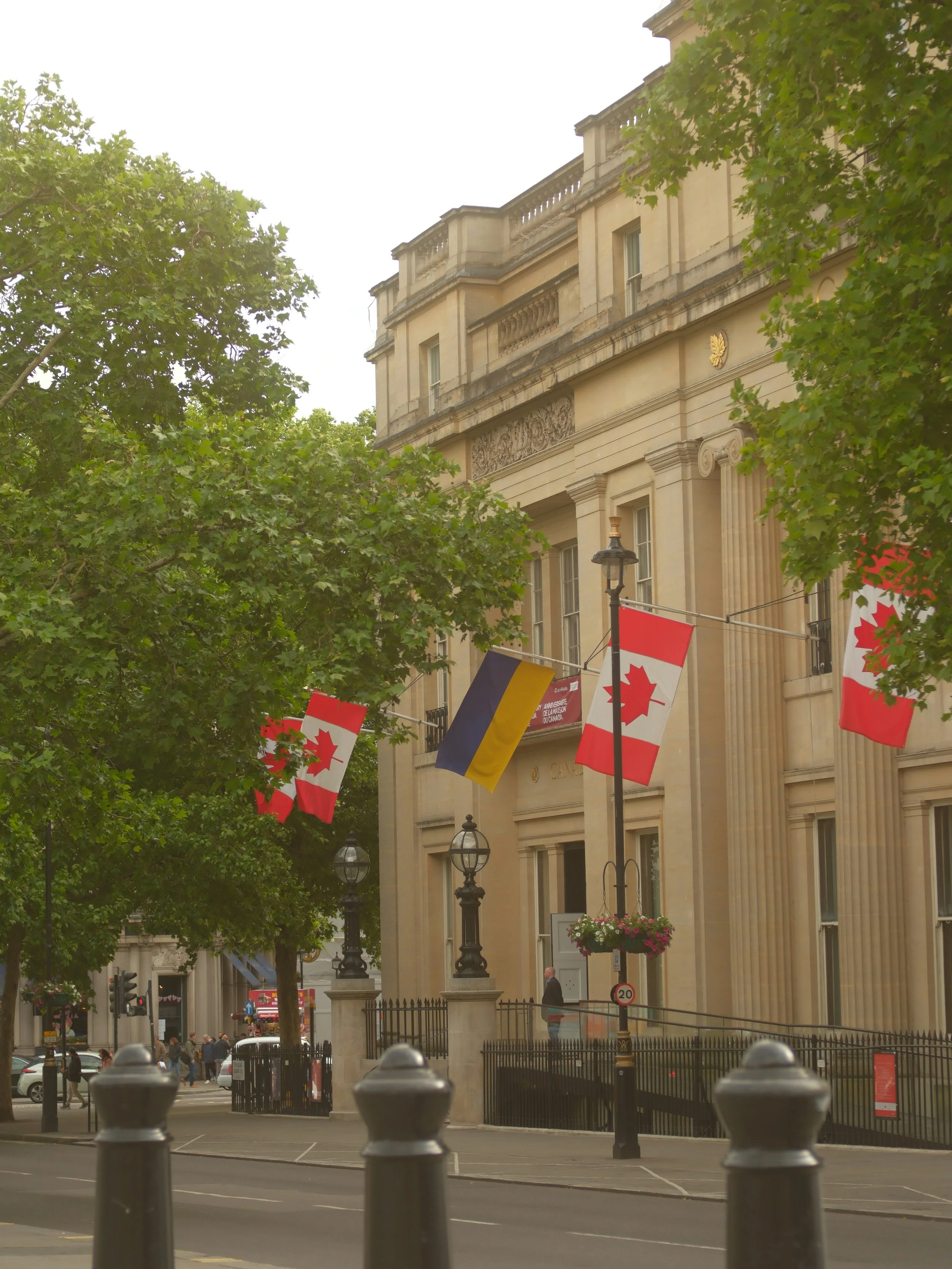 Street view of a historic building with flags of Canada and Belgium, trees lining the sidewalk, and street lamps.