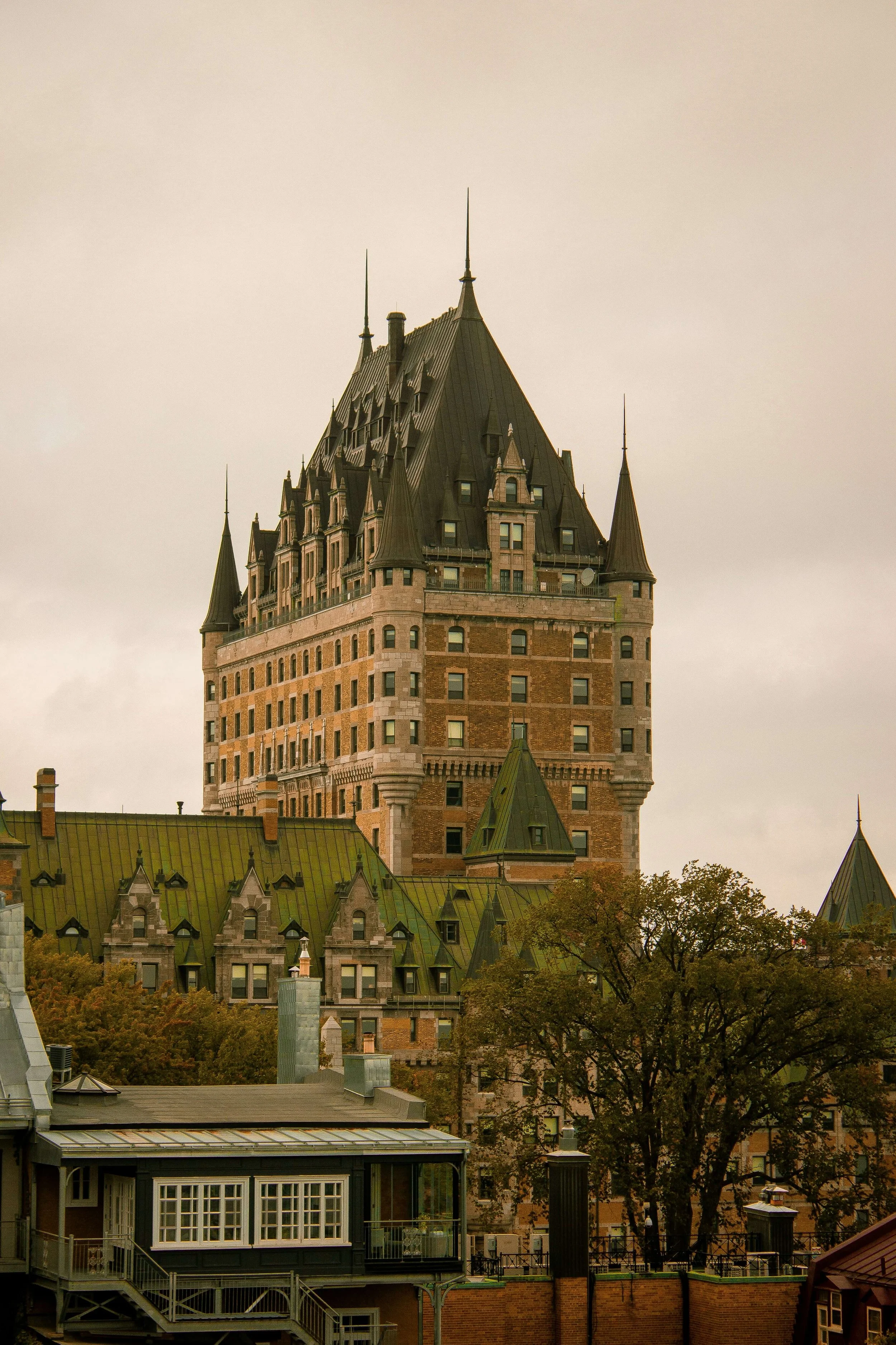 View of a historic castle-like building with a tall, pointed roof and turrets, surrounded by trees and other buildings.