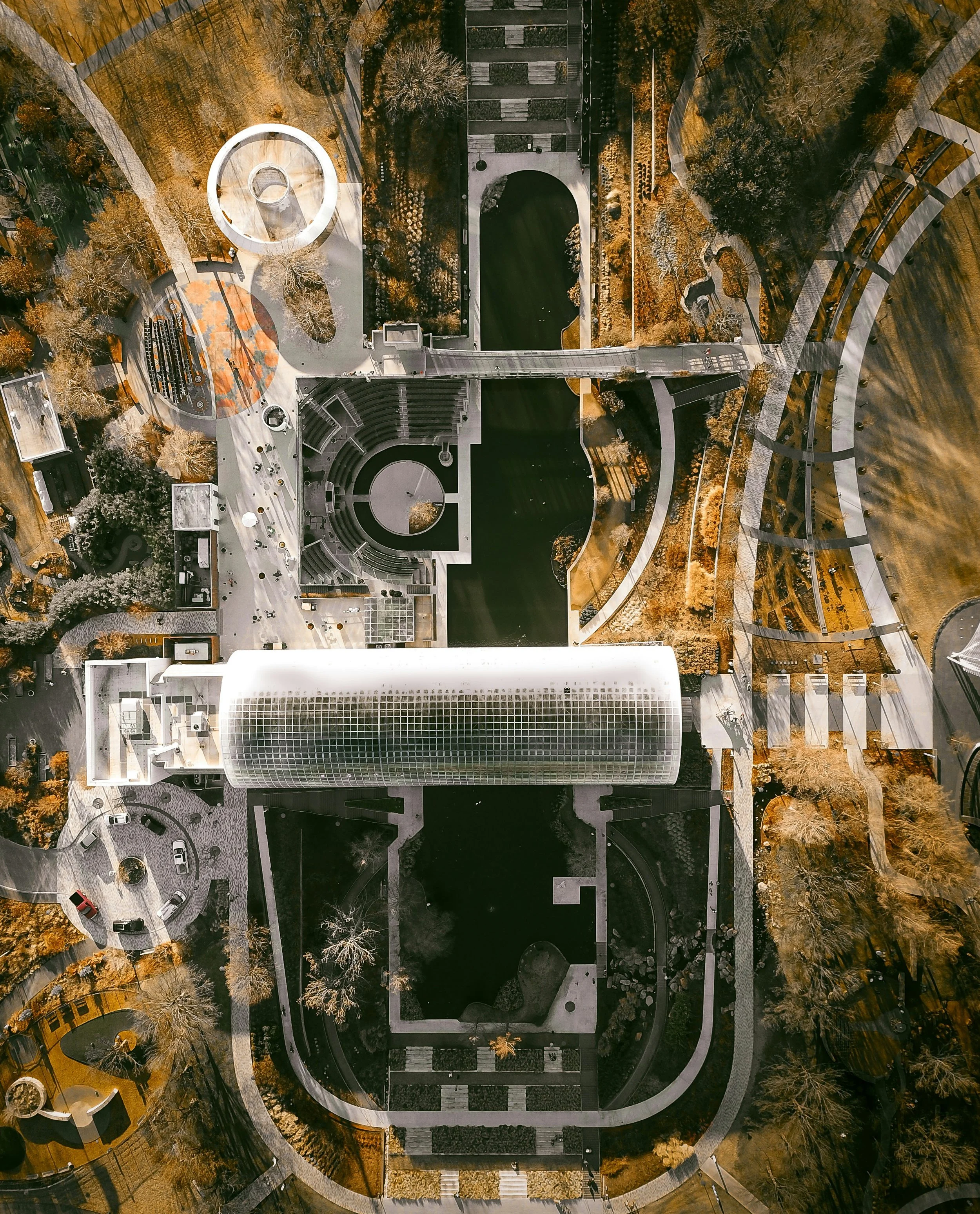 Aerial view of a modern urban park with winding pathways, trees with fall-colored leaves, and various architectural features including a cylindrical building with a glass exterior, a circular amphitheater, and bridges over a body of water.