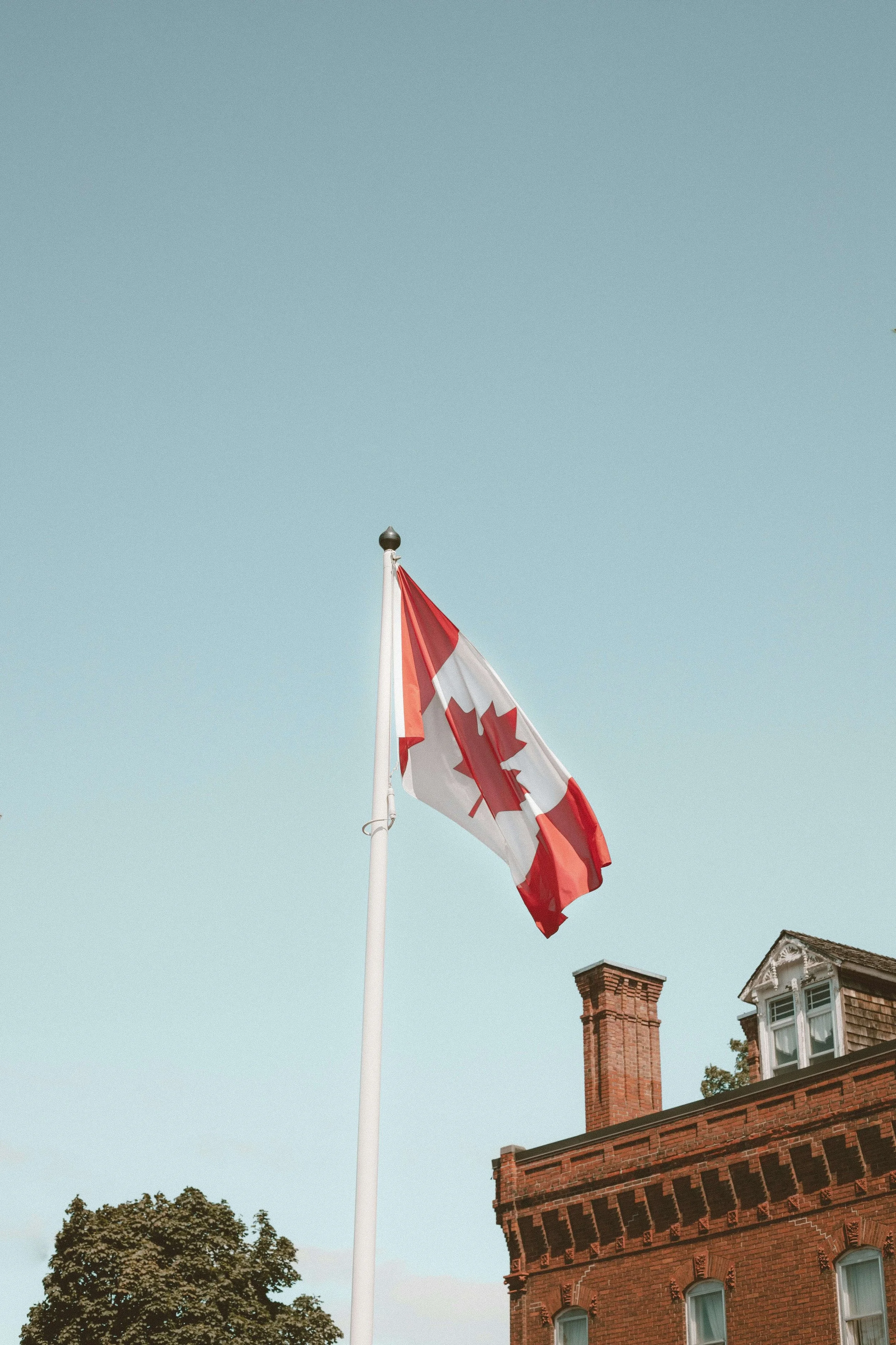 Canadian flag flying on a white pole in front of a brick building with a chimney and a tree.