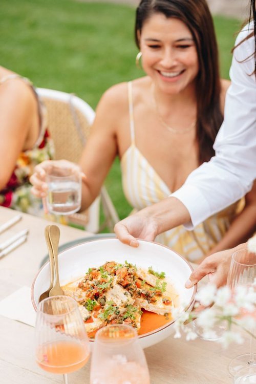 A woman in a yellow and white striped dress smiling while sitting at an outdoor gathering, with a server presenting a dish of food on a white plate.