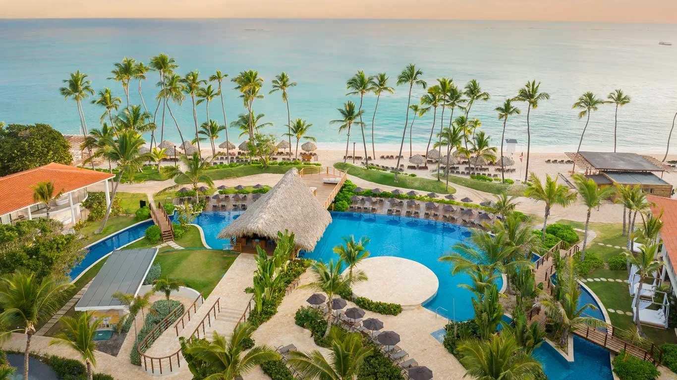 Aerial view of a tropical beachfront resort with a large swimming pool, lush palm trees, beach chairs, and ocean in the background at sunset.
