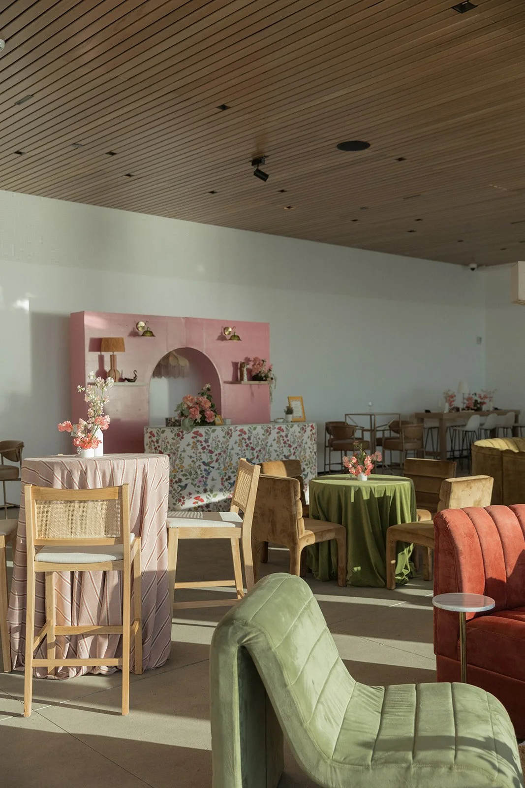 Interior of a decorated event space with pastel-colored furniture and floral decor, including round tables with flowers, chairs, and a pink floral backdrop.