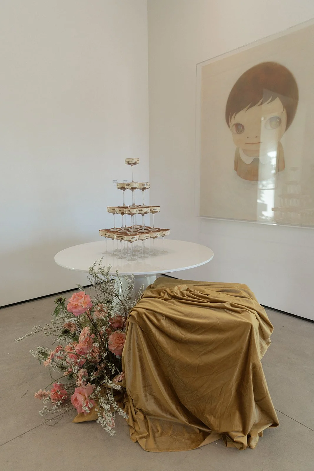 A champagne tower made of stacked glasses on a white round table, decorated with pink flowers and a gold cloth draped over an object, with a large framed cartoon portrait on the wall.