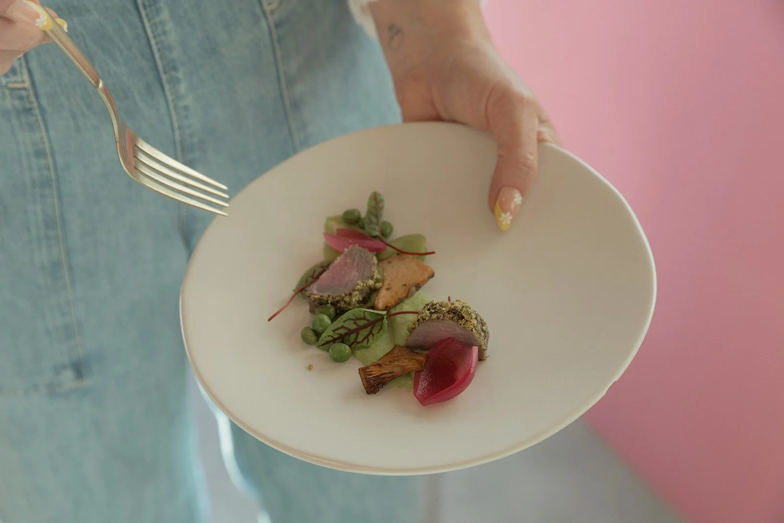 Person holding a white plate with gourmet food, including sliced vegetables and garnishes, standing against a pink background.