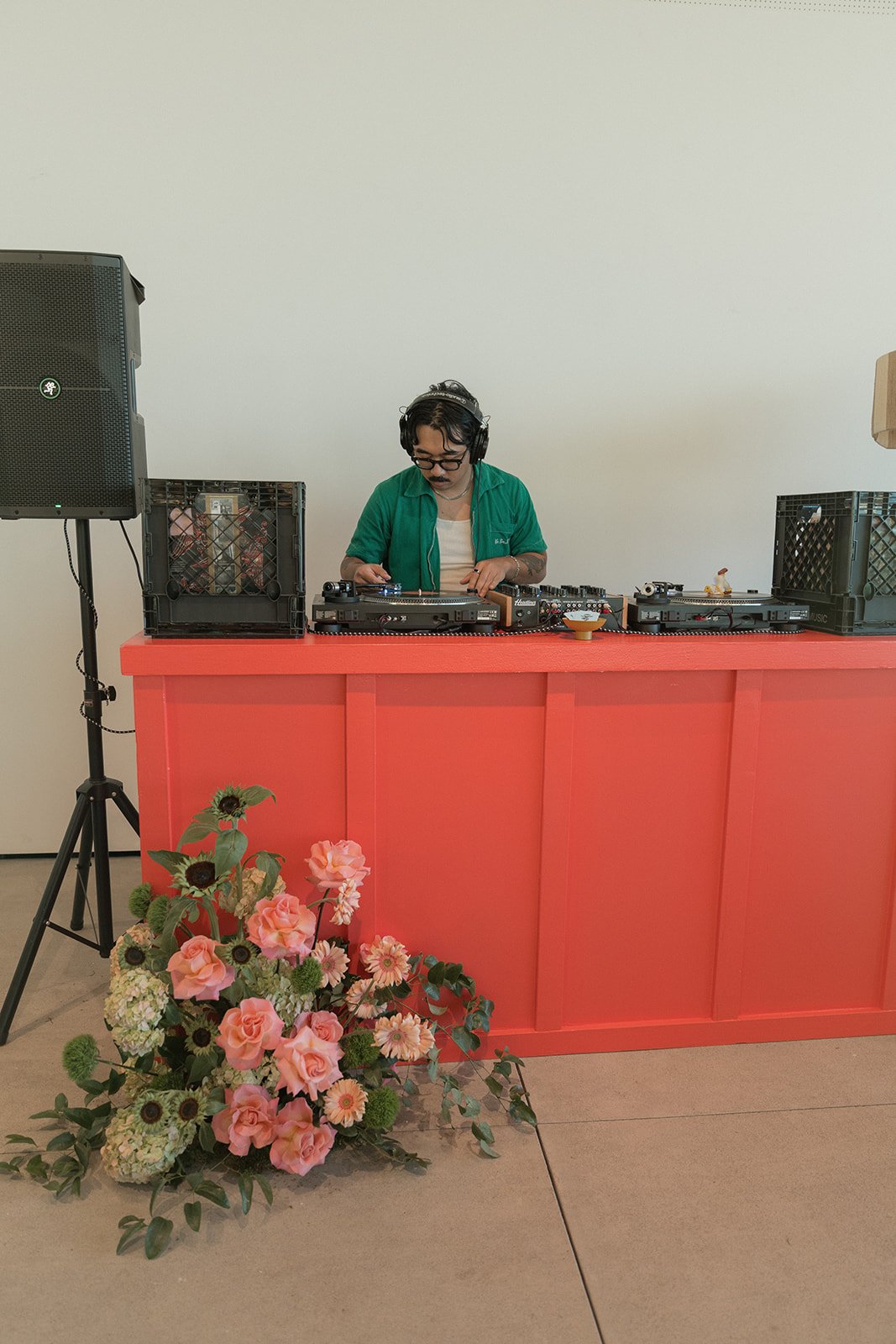 A DJ with headphones and glasses performing at a coral-colored bar with a floral arrangement in front, against a plain white wall.