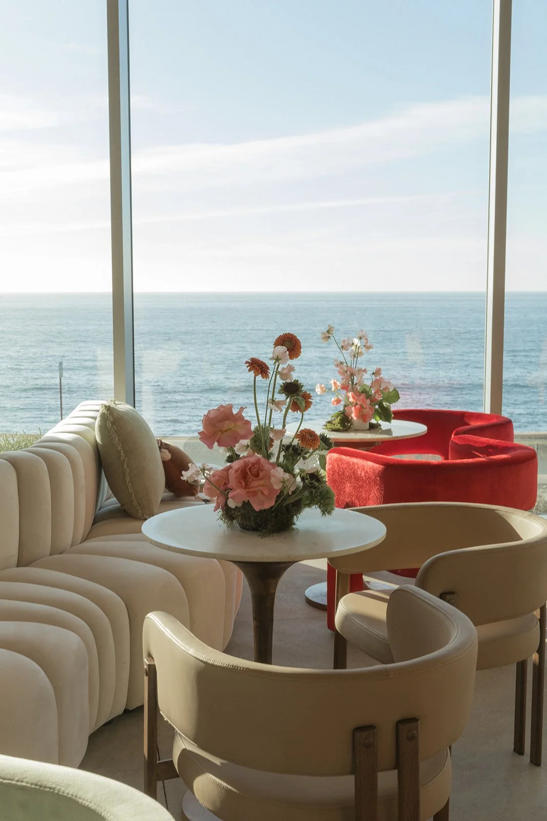 Indoor seating area with beige velvet sofas and chairs, floral arrangements with pink and orange flowers, large windows overlooking the ocean, and a bright sky.