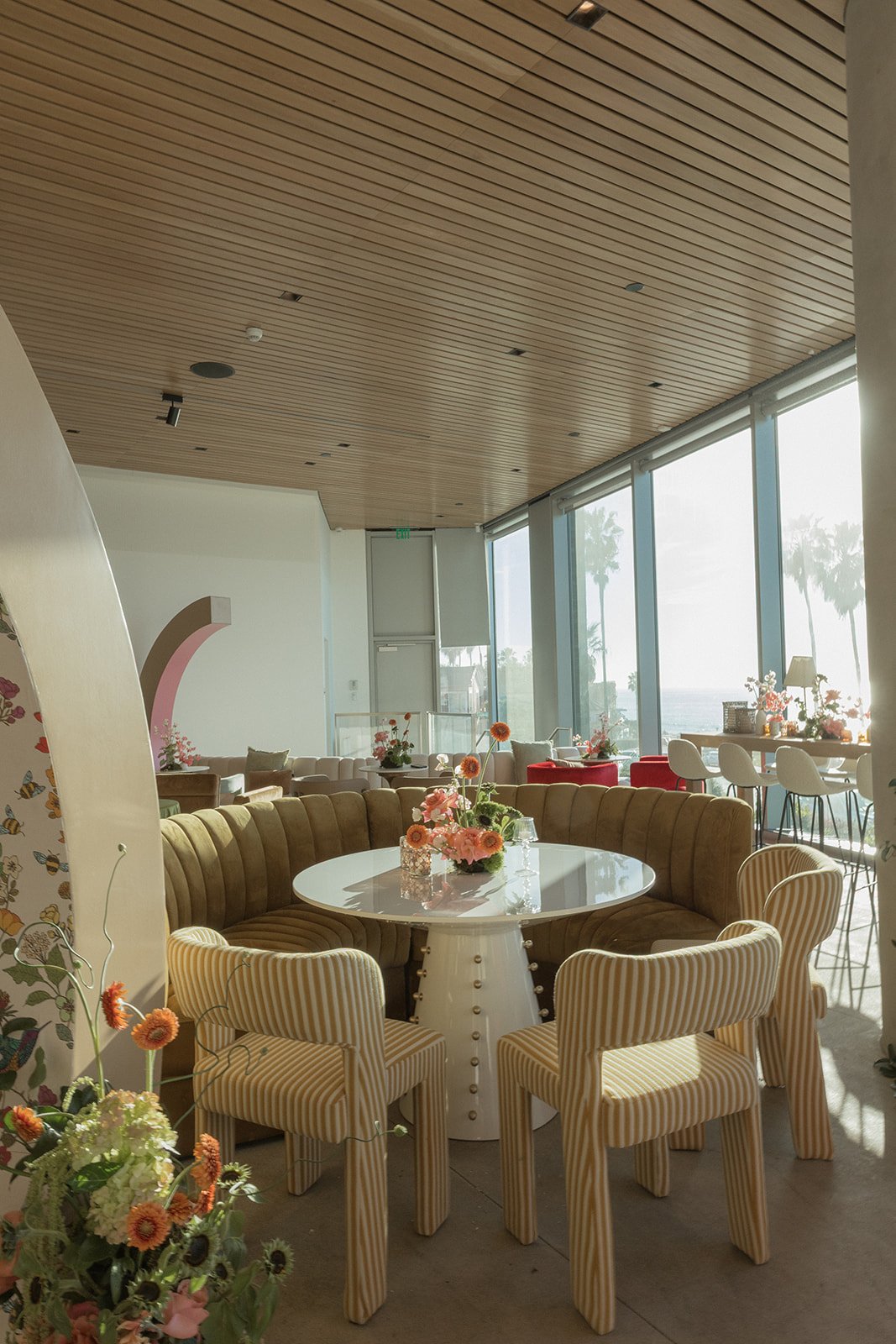 Interior of a bright, modern cafe or restaurant with large windows, a round table with a flower centerpiece, striped chairs, and a wooden ceiling.