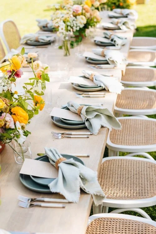 A long outdoor dining table set with plates, napkins, forks, and colorful flower arrangements for a gathering or celebration.