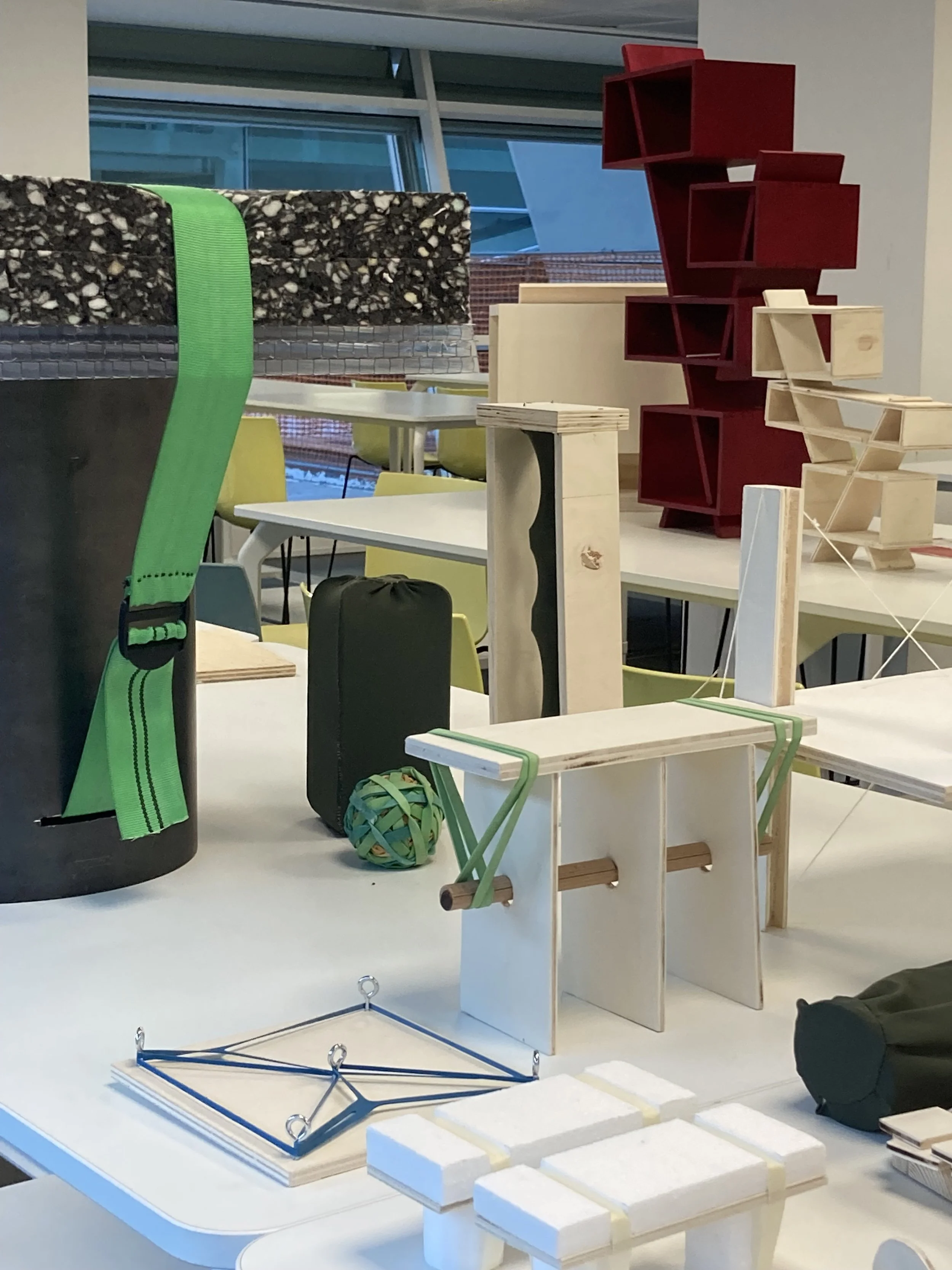 Modelli di mobili innovativi e giochi di design su un tavolo, con scaffali rossi, strutture in legno e oggetti colorati in un ambiente di prototype o laboratorio.