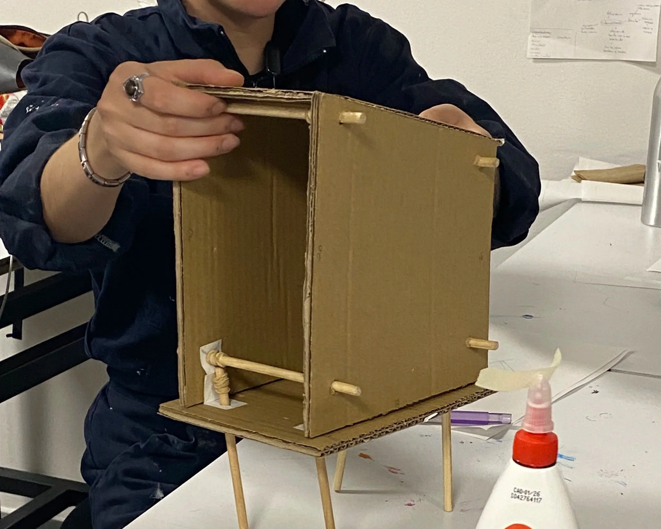 Persona che lavora su una scatola di cartone assemblata con stecchi di legno, in un ambiente di lavoro o di laboratorio.