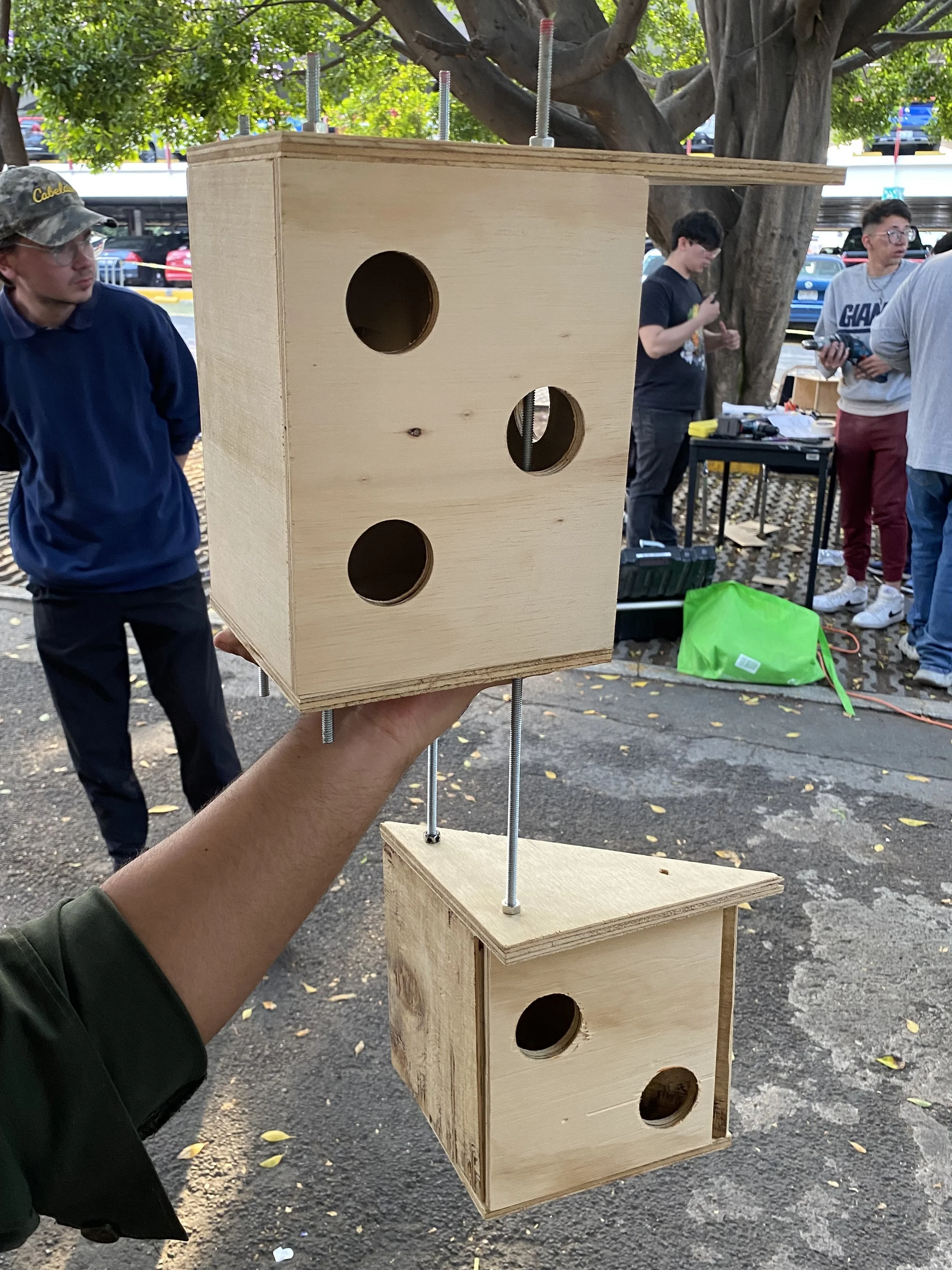 Una persona sta tenendo in mano due casette per uccelli di legno con fori, con persone e attrezzature sullo sfondo in un'area all'aperto.