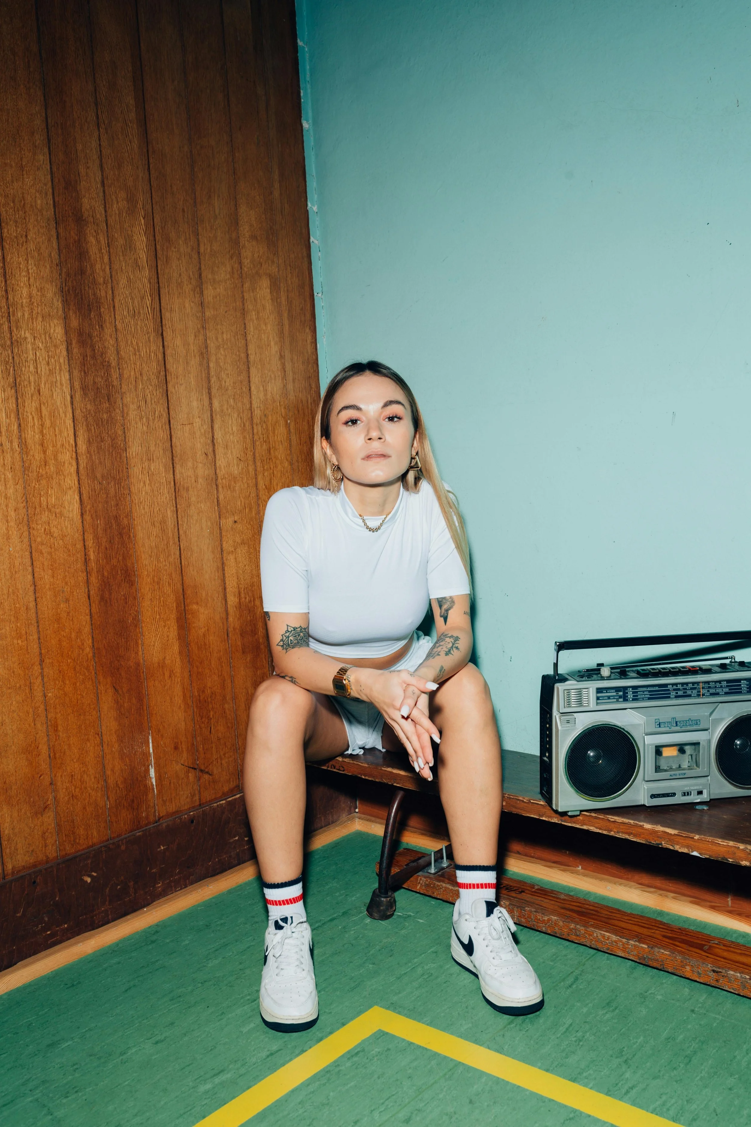 A young woman with tattoos wearing a white t-shirt, shorts, and sneakers sitting on a wooden bench in a room with green carpet and wood-paneled wall, next to a vintage boombox.