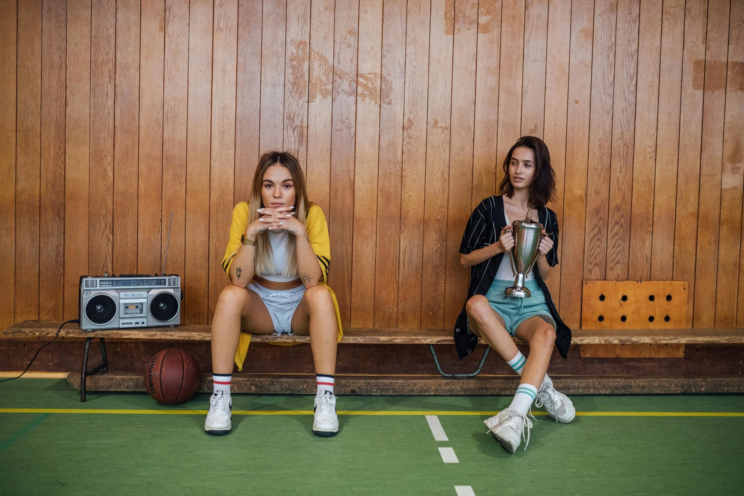 Two young women sitting on a wooden bench in a gymnasium, one holding a trophy and the other sitting with a boombox and a basketball nearby.