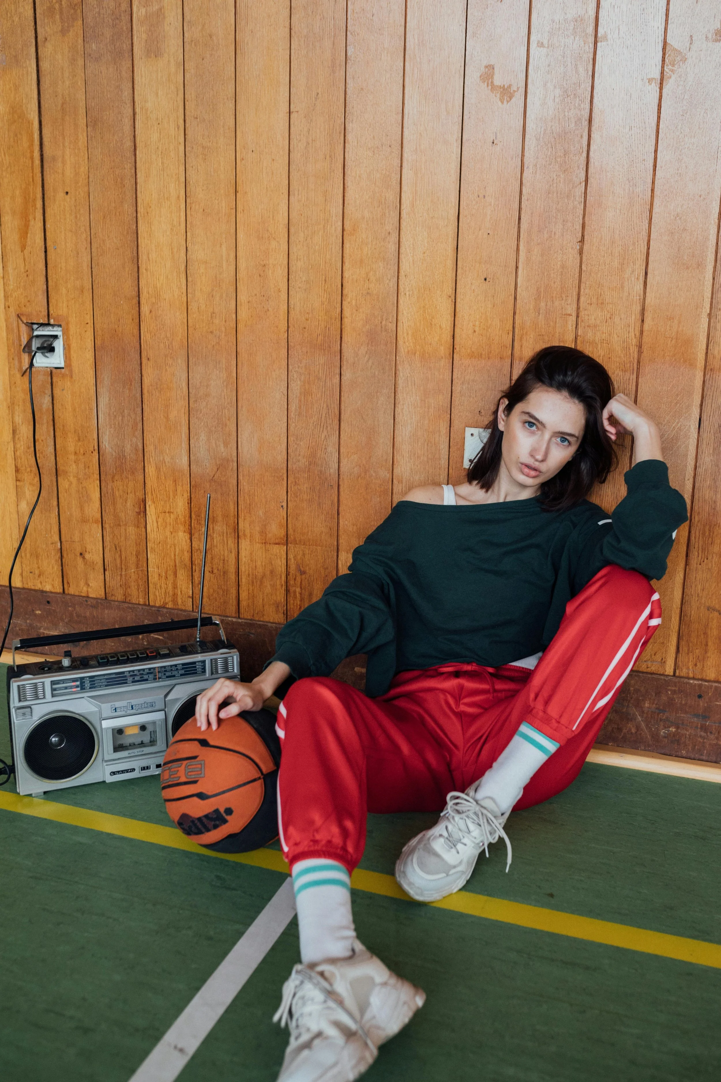 A young woman with dark hair in a gymnasium, sitting on the floor against a wooden wall. She wears a dark green sweatshirt, red athletic pants, and white sneakers, with one hand resting on a basketball. There is a retro boombox and a power outlet on the wall behind her.