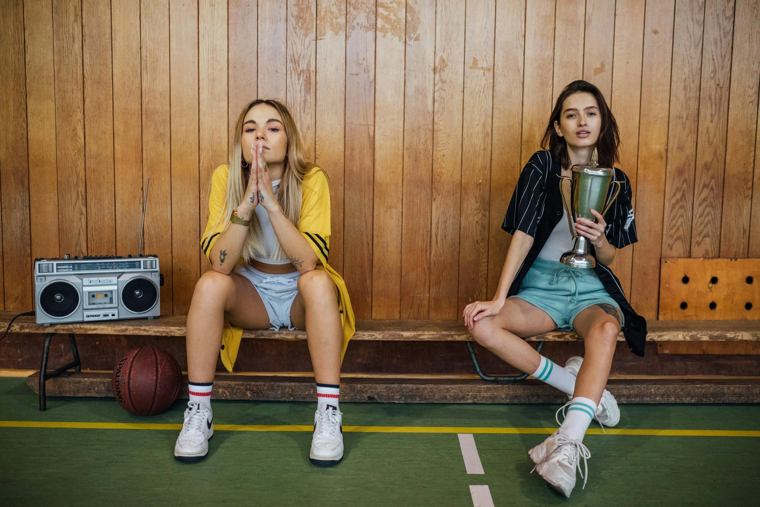 Two young women sitting on a wooden bench inside a gymnasium with wood-paneled walls. One on the left is wearing a yellow jacket, white shorts, sneakers, and has tattoos, with hands clasped in front of her face. The other on the right, wearing a black and white striped shirt, teal shorts, and white sneakers, is holding a large trophy. There is a boombox and a basketball on the floor in front of them.
