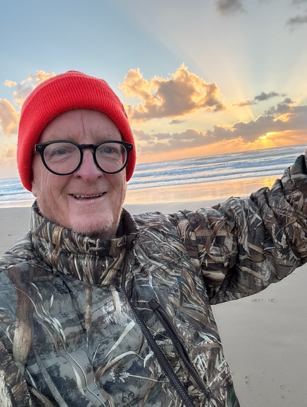 Man taking a selfie on the beach at sunset, wearing a red beanie, glasses, and a camouflage jacket.