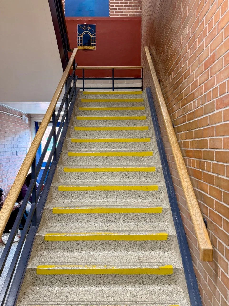 Staircase with yellow safety strips and wooden handrails, brick wall to the right, blue railing to the left, and a poster at the top of the stairs.