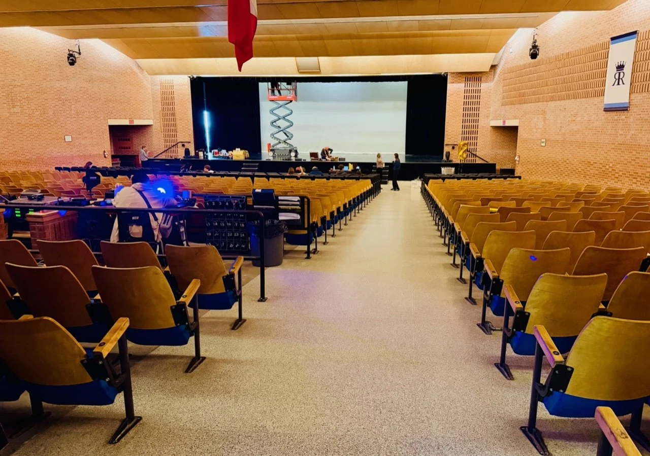Empty auditorium or theater with rows of yellow seats, a stage at the front, and crew members preparing behind the scenes.