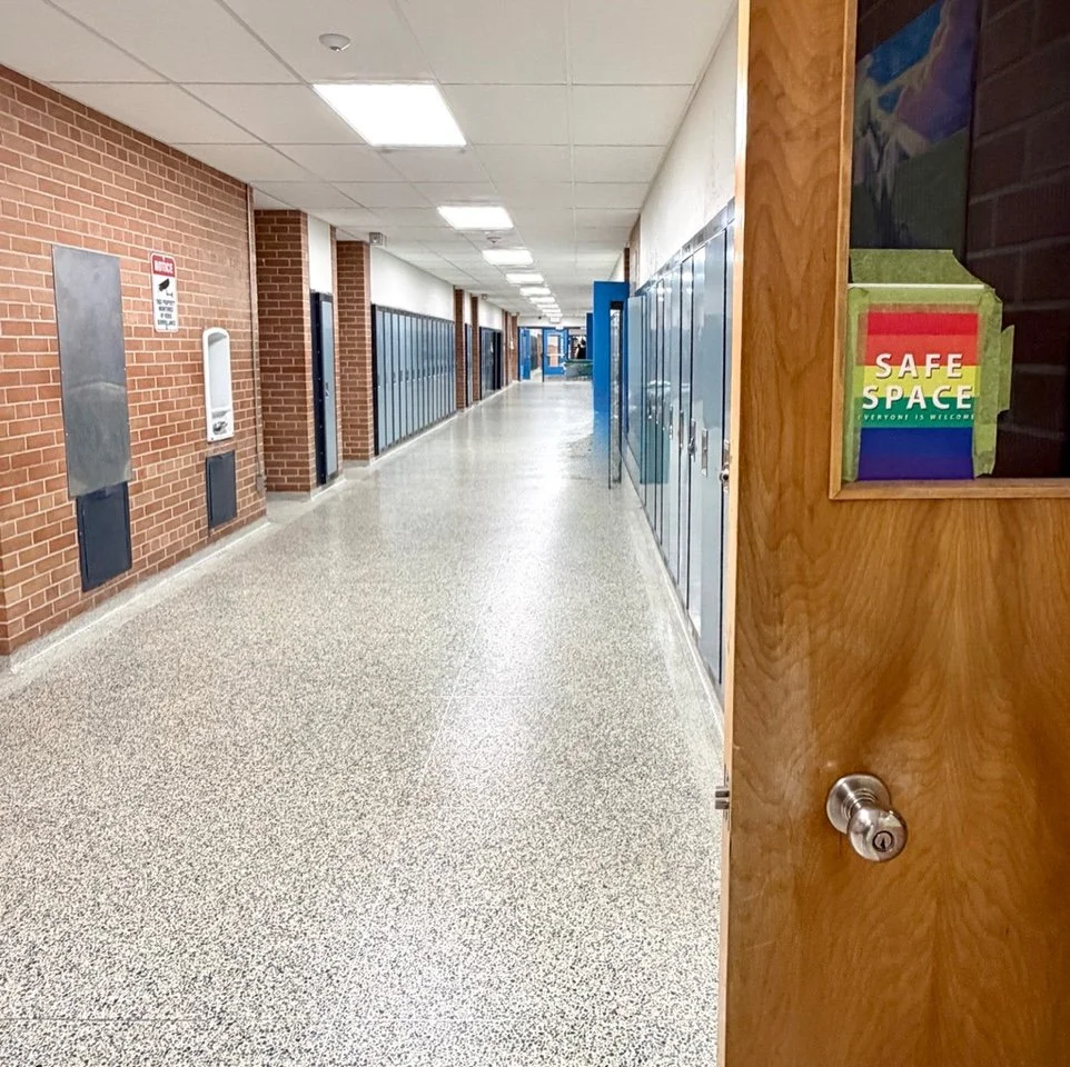 A school hallway with blue lockers on the right and brick walls on the left. The hallway has a speckled floor, a white ceiling with rectangular fluorescent lights, and a wooden door with a 'Safe Space' sign on it.