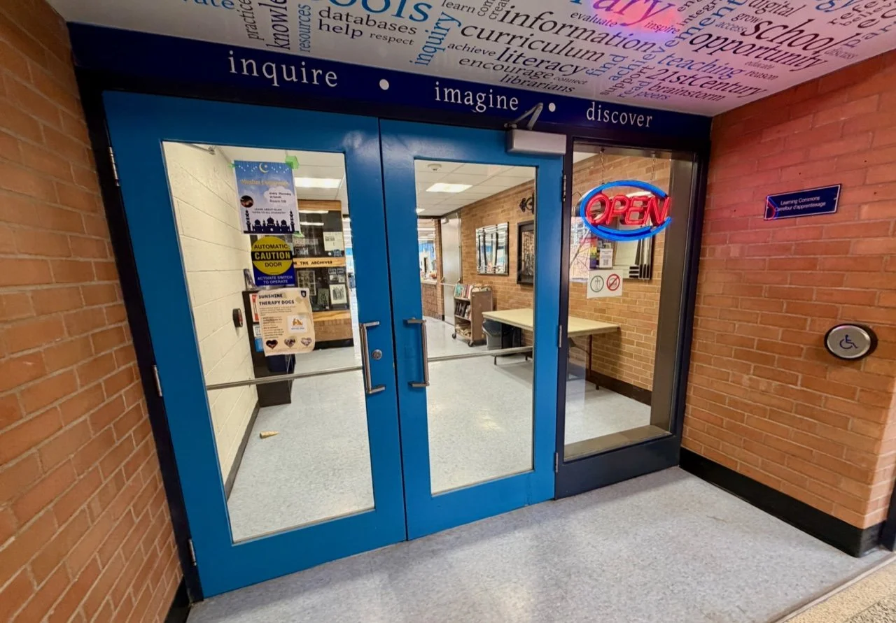 Blue double doors with a glass window and an illuminated red-and-blue 'OPEN' sign, leading into a library or learning center with brick walls and informational posters inside.