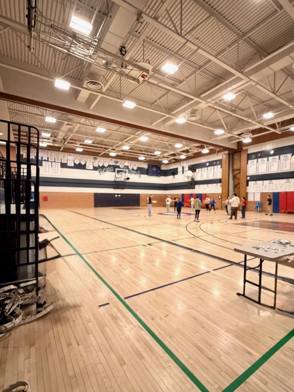 School gymnasium with basketball court and several people standing and talking, with tables and equipment on the sides.