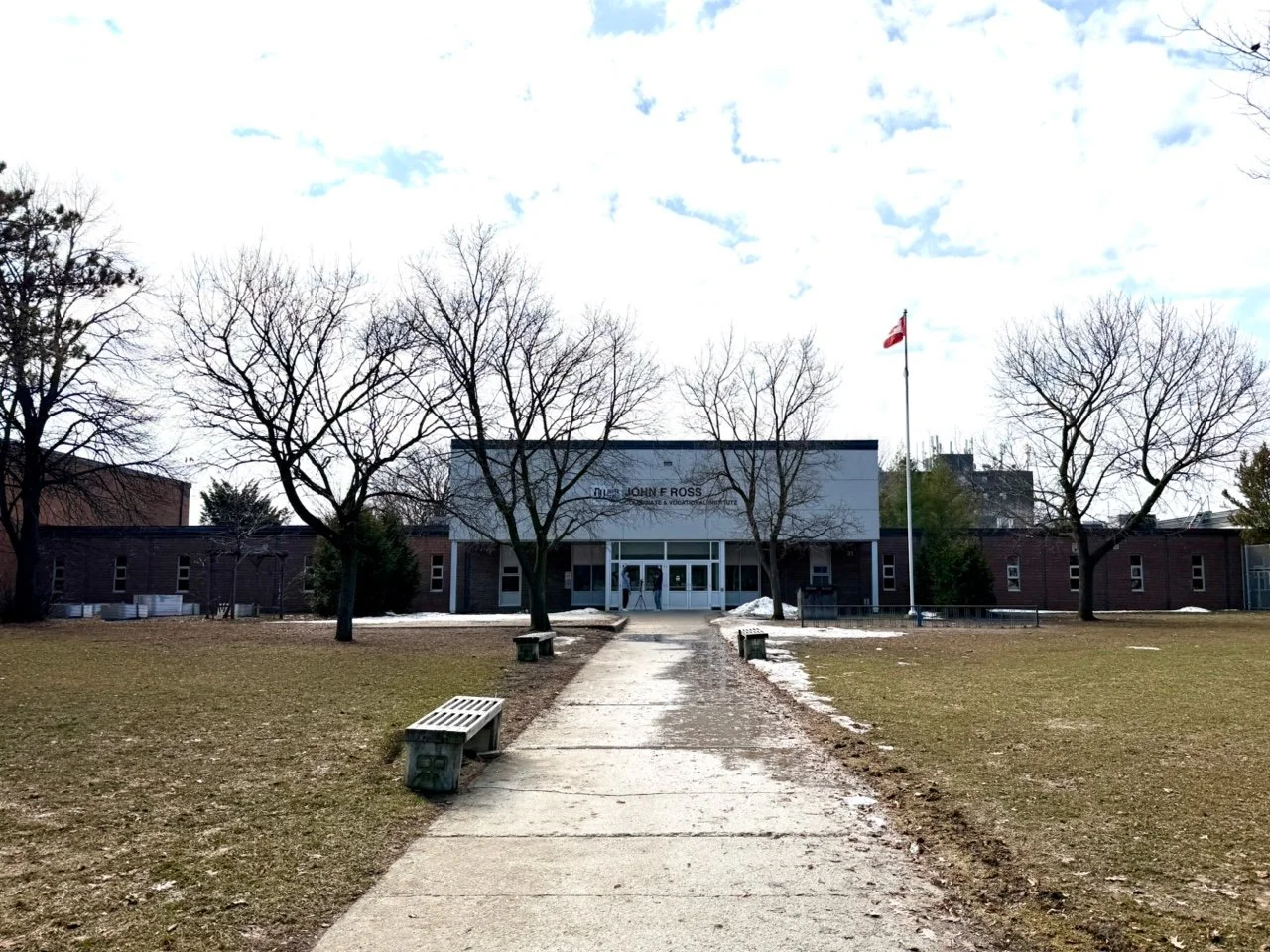A school building with a sign that reads 'John F. Ross' in a park-like area with leafless trees, a flagpole with a flag, benches, and patches of snow on the ground.