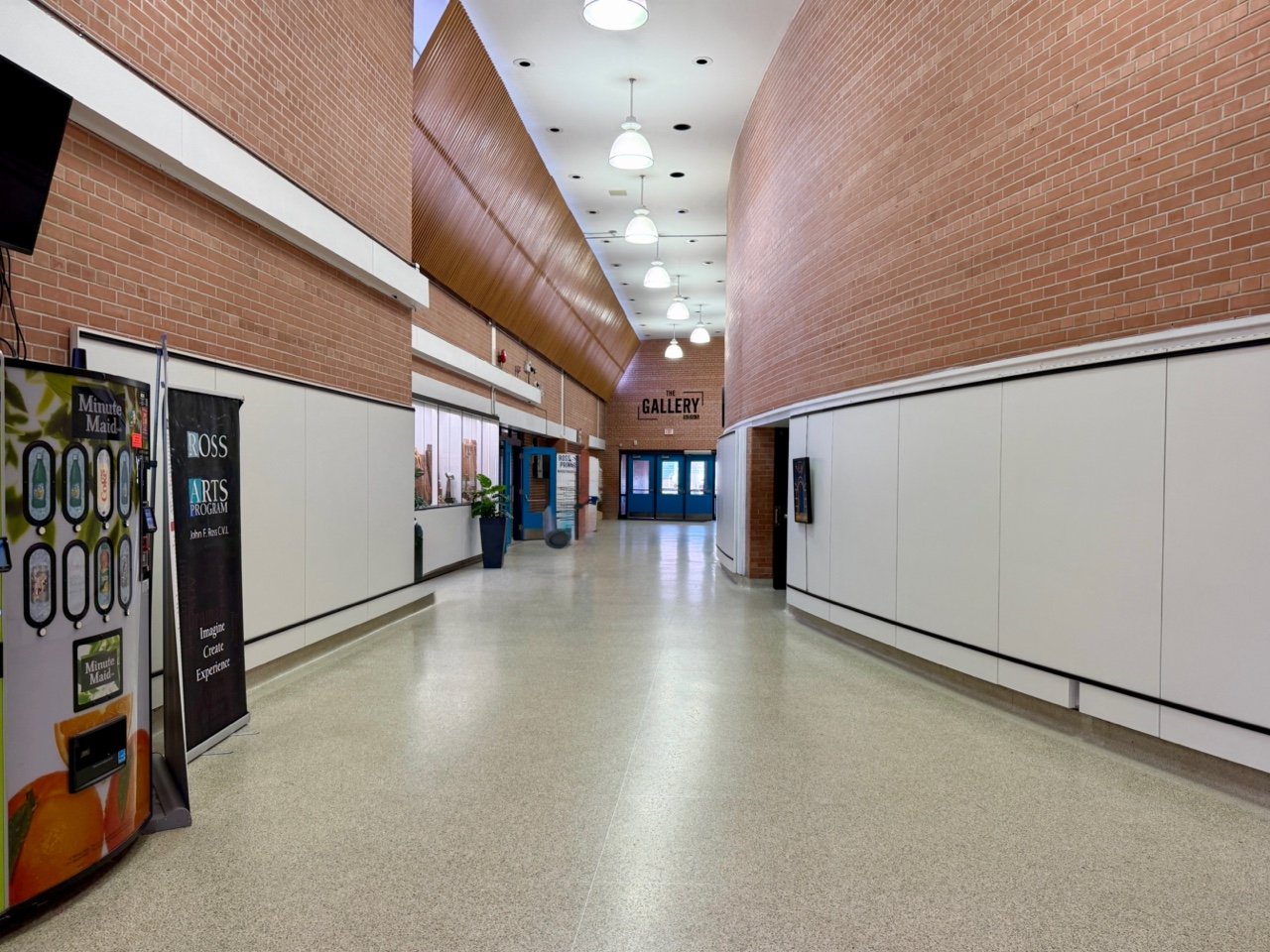 Empty hallway in an art gallery with white walls, brick accents, and a curved design. Doors and signs are visible, with a vending machine on the left and ceiling lights overhead.