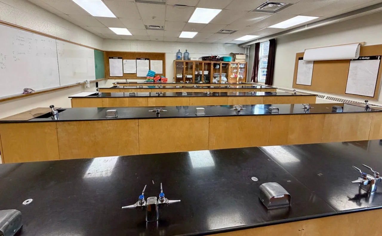 Empty science classroom with black lab tables, whiteboards, and cabinets filled with supplies, lit by overhead fluorescent lights.