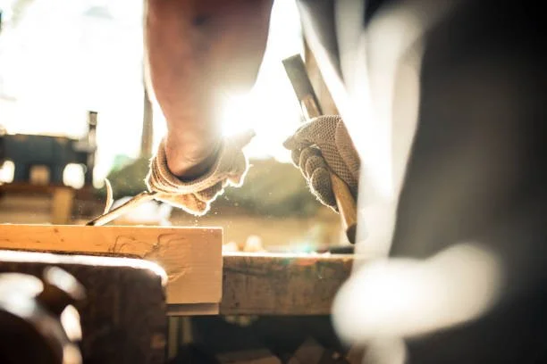 Two people woodworking, one holding a chisel and the other steadying wood, with sunlight in the background.