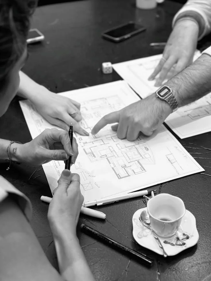 Two people discussing architectural plans or blueprints on a table, with writing utensils, a coffee cup, a smartphone, and a small die nearby.