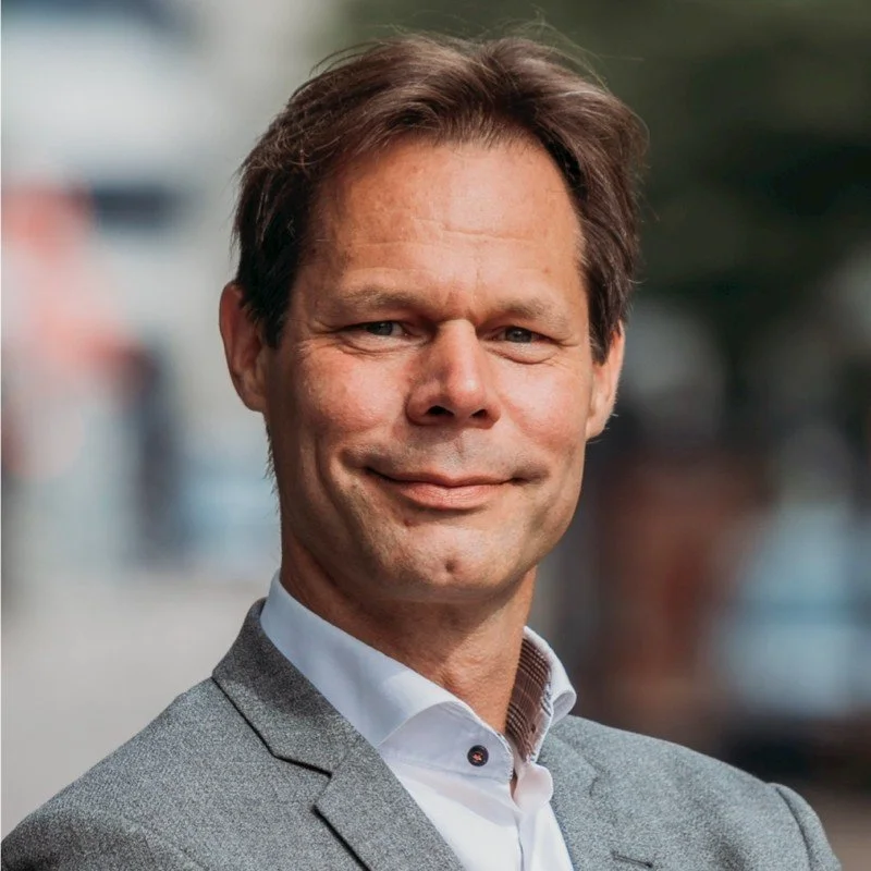 Een man in een grijze jas en witte blouse, glimlachend met een stedelijke achtergrond.