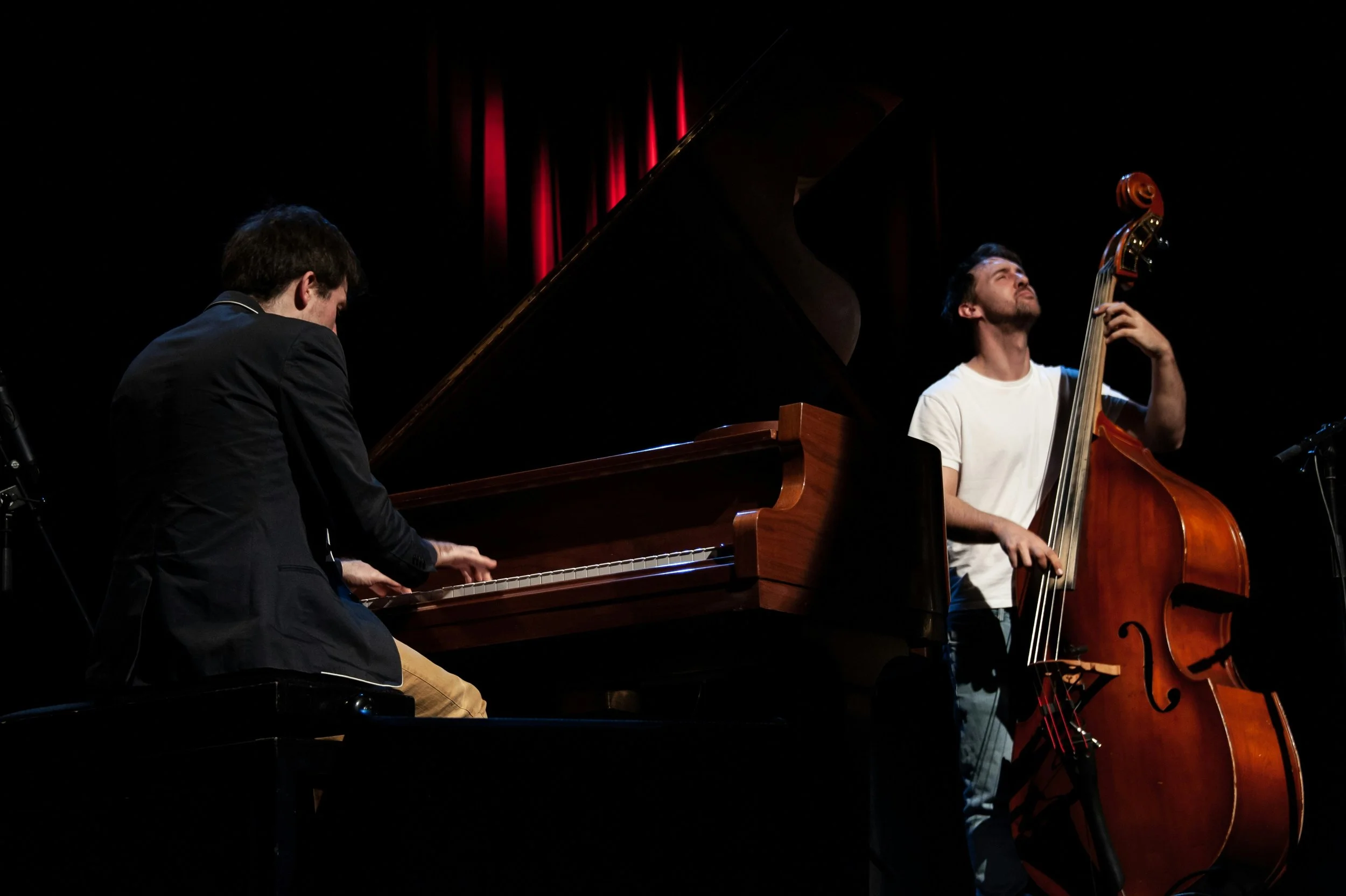 Twee muzikanten spelen jazzmuziek: een man op piano en een man op contrabas voor een zwart podium met rode verlichting.