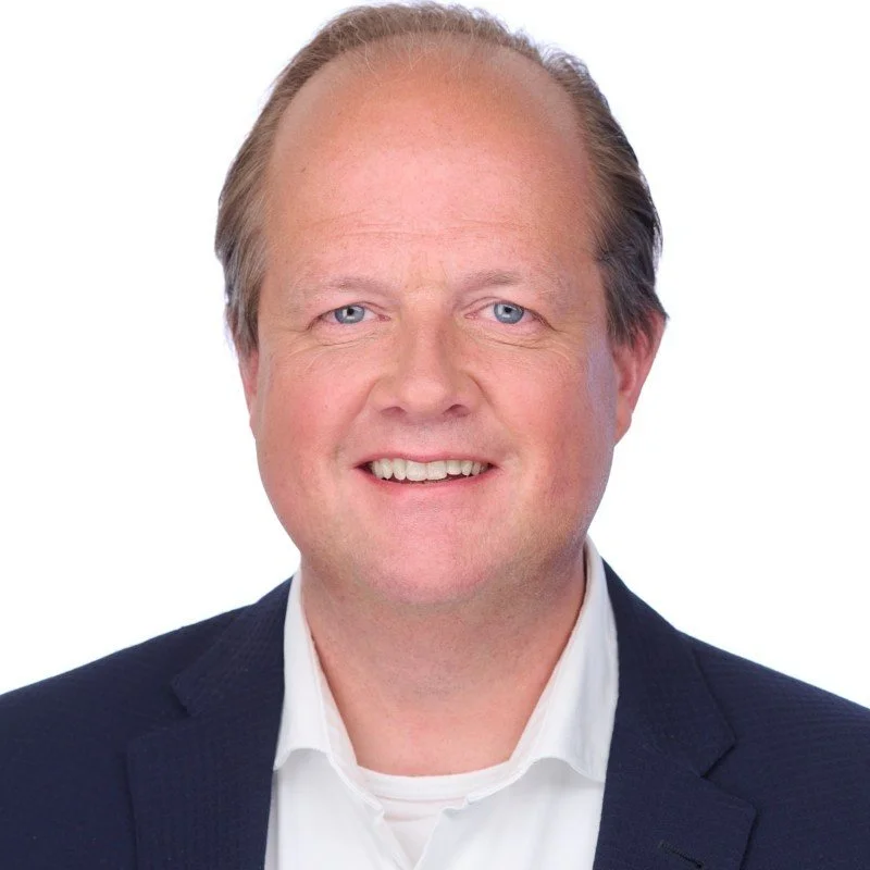 Een man met lichte huid, blond haar en blauwe ogen, gekleed in een donkerblauw pak en witte overhemd, lachend naar de camera.