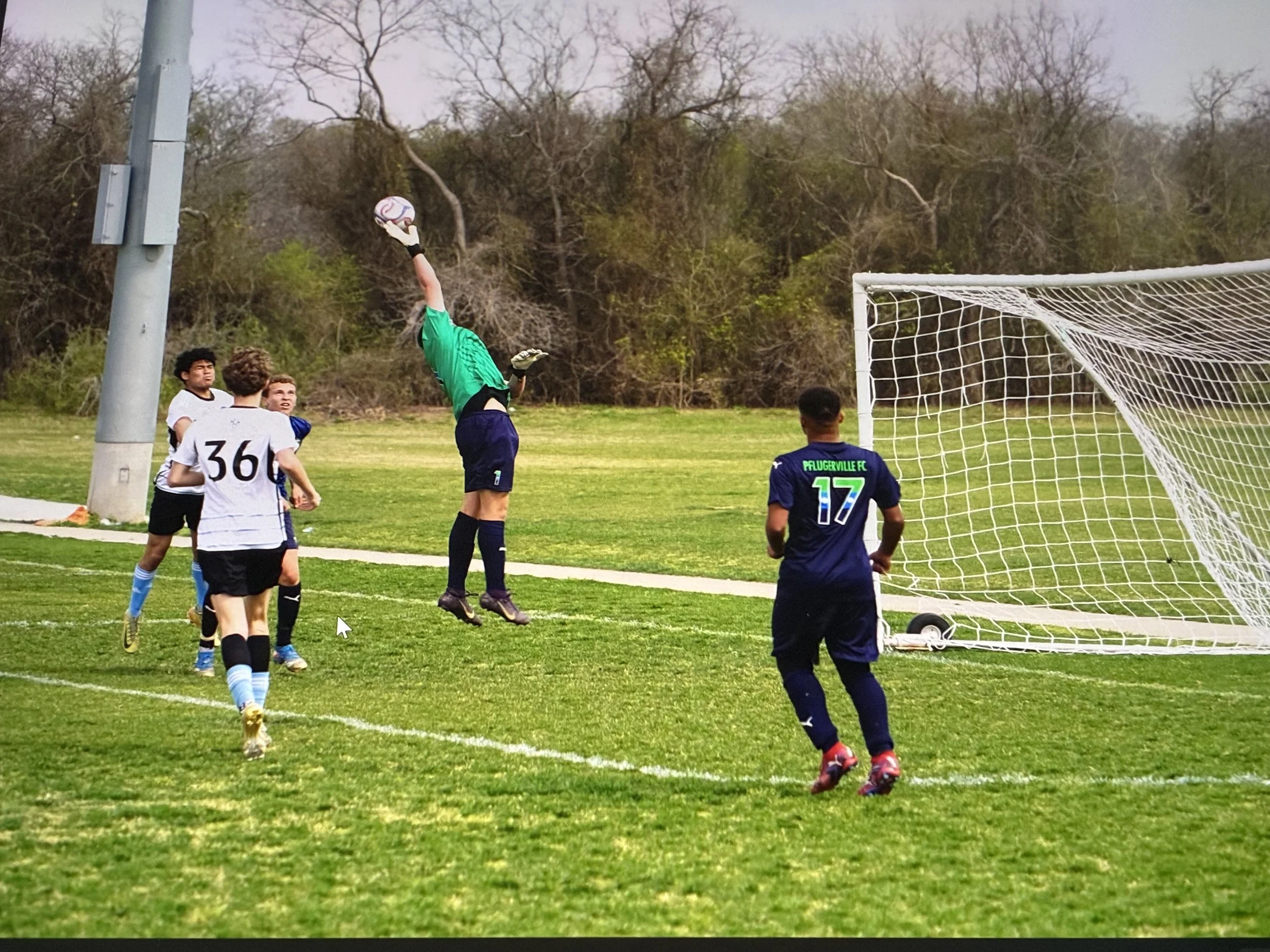 A youth soccer game with players near the goalpost. One player in a green goalkeeper jersey is jumping to catch or block a soccer ball, while others are observing or preparing for the play. Tree-filled background and an outdoor field.